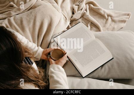 unrecognizable Woman sitting on the bed reading a book and drinking coffee Stock Photo