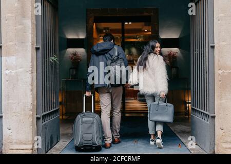 Back view of happy stylish young couple in love with suitcase and bags holding hands and entering modern city hotel during travel while Woman looking at camera over shoulder Stock Photo