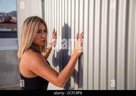 Back view from above of stylish blond haired female in sportive clothes looking away while leaning on wall stretching legs on rooftop Stock Photo