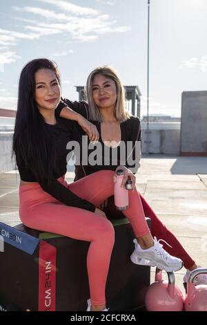 Pretty athletic female friends in active wear smiling while looking at camera after workout on roof of building Stock Photo