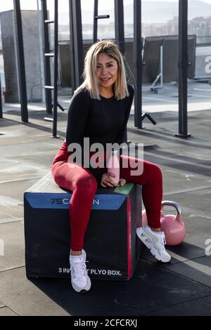 Confident blond haired female athlete in stylish active wear sitting and looking at camera with smile on urban background Stock Photo