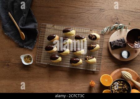 Top view of homemade freshly baked Madeleine cookies dipped on chocolate and sprinkled with nuts on wooden kitchen table with recipe ingredients and utensils Stock Photo