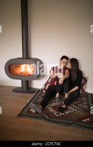 Happy couple in casual clothes embracing each other sitting on floor near fireplace on colorful carpet against white wall at home Stock Photo