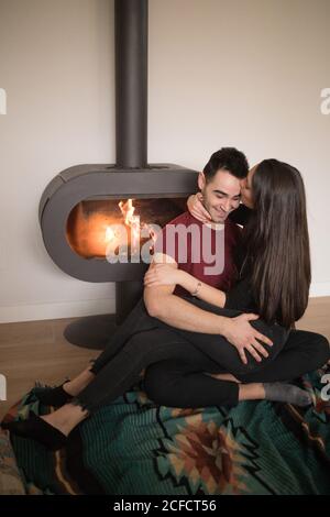 Happy couple in casual clothes embracing each other sitting on floor near fireplace on colorful carpet against white wall at home Stock Photo