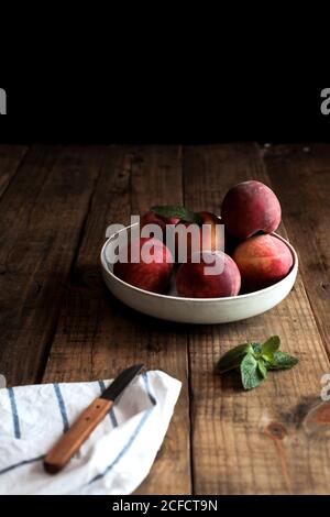 Appetizing bright peaches with leaves in white plate on wooden table and knife on cloth napkin Stock Photo