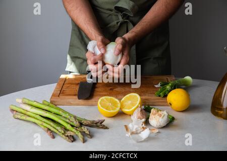 Crop anonymous man peeling onion while standing at table with fresh ingredients for healthy dinner in home kitchen Stock Photo