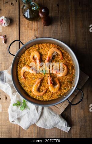 High angle of big metal pan of while appetizing paella with roasted shrimps on rustic table in composition with vintage bottle of oil and towel in kitchen at home Stock Photo
