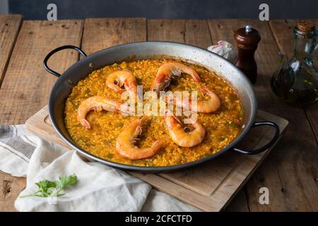High angle of big metal pan of while appetizing paella with roasted shrimps on rustic table in composition with vintage bottle of oil and towel in kitchen at home Stock Photo