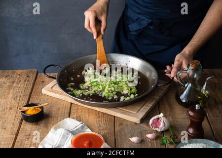 High angle of crop cook in dark apron stirring with wooden spatula mix of chopped green chili pepper and onion on big metal pan while adding oil before firing in contemporary kitchen at home Stock Photo