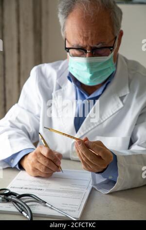 Male general practitioner wearing protective mask and medical gown sitting at table in hospital and taking notes in report about diagnosis of patient while working during quarantine Stock Photo