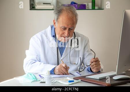 Male general practitioner wearing medical gown sitting at table in hospital and taking notes in report about diagnosis of patient while working during quarantine Stock Photo