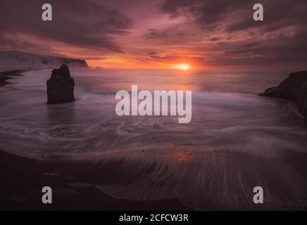 Sun setting over horizon and reflecting in sea water at Icelandic snow covered rocky coast under beautiful cloudy sky Stock Photo