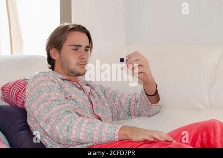 Sick looking young man checking an electronic thermometer while lying on a sofa on an out of focus background. Healthcare and sickness concept. Stock Photo