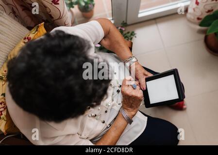From above of elderly gray haired Woman in shirt and glasses sitting on armchair and reading e-book in apartment Stock Photo