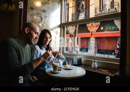 Handsome bald man and attractive Woman enjoying coffee and tea while sitting at small table near window during date in cozy cafe Stock Photo