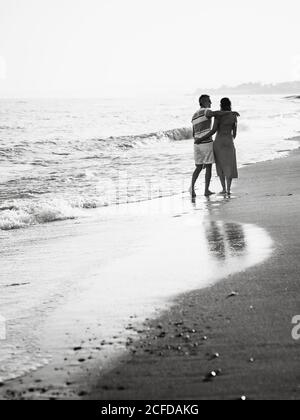 Back view of barefoot man and Woman holding hands and carrying shoes while walking on sandy beach towards waving sea on resort Stock Photo