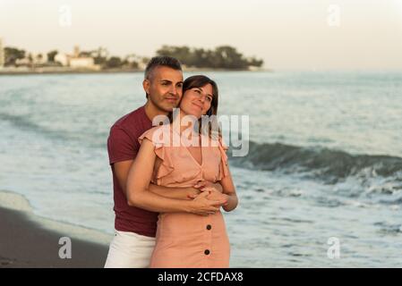 Adult man hugging Woman from behind and looking away while standing on beach near waving sea and resting together Stock Photo