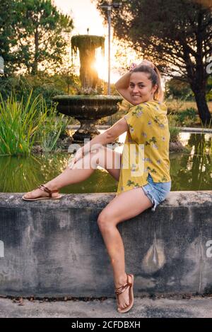 Charming smiling young Woman in casual wear sitting on fountain at sunset posing and looking at camera in back lit Stock Photo