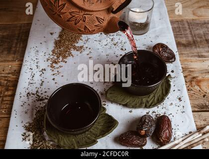 Serving fragrant tasty tea in cup clay teapot and sweet dates on white tray decorated with tea leaves on wooden background Stock Photo