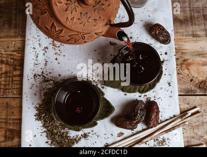 Serving fragrant tasty tea in cup clay teapot and sweet dates on white tray decorated with tea leaves on wooden background Stock Photo
