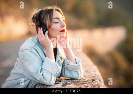 Side view of satisfied young lady in stylish casual wear and red lips leaning elbow on rocky fence with closed eyes while listening music with headphones Stock Photo