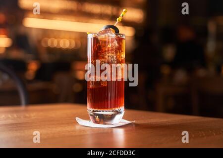 Highball glass with red alcohol cocktail with ice cubes decorated with stick with black olive on wooden counter Stock Photo