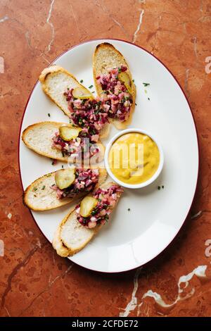 Top view of roasted slices of bread with colorful salad and rounded pieces of salted cucumbers on oval white plate with yellow sauce Stock Photo