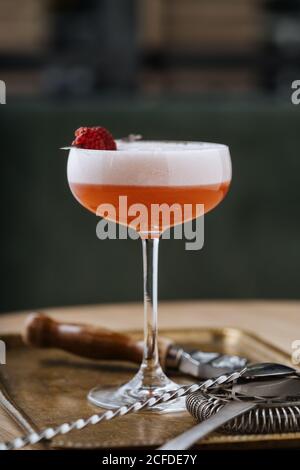 Colorful red alcohol cocktail in stylish glass on table with bar spoon and strainer in restaurant Stock Photo