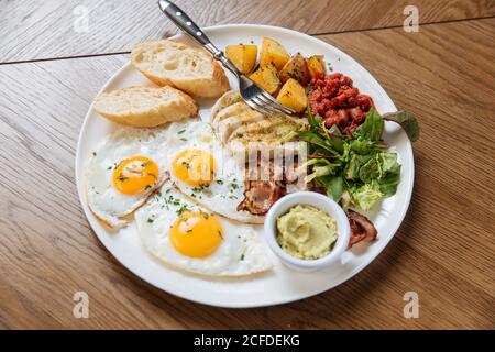 Top view of served English breakfast with fried eggs and bacon with toasts and vegetables on wooden table Stock Photo