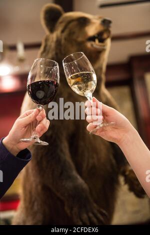 Crop couple toasting with glasses of red and white wine while having romantic dinner in hunter style restaurant with bear scarecrow in background Stock Photo