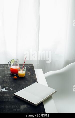 From above of jar and glass with red and orange fruit drinks placed with book on table near window in light room Stock Photo