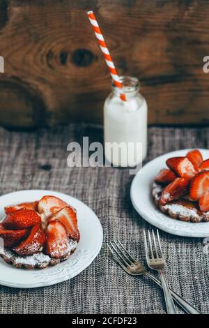 From above of delicious homemade waffles with ripe strawberry and sugar powder served on white plates on table with milk drink in glass bottle Stock Photo