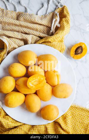 Top view of white plate with fresh yellow ripe apricots placed on yellow cloth on white marble table with cut in half apricot Stock Photo