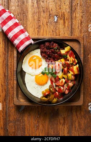 Top view of fried eggs with pieces of meat and vegetables in pan on wooden table Stock Photo