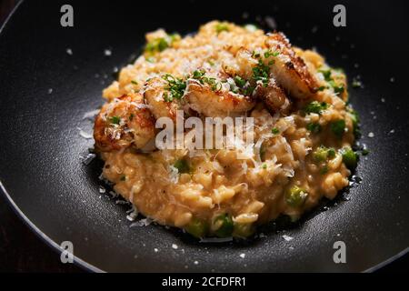 From above of rice in golden sauce decorated with fried shrimps grated cheese and green herbs on stylish black plate Stock Photo