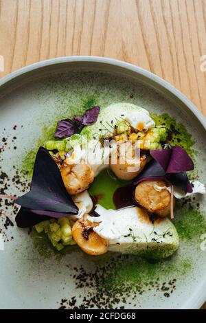 Steamed white fish steaks with shrimps and purple basil leaves on white plate garnished with green matcha powder on wooden table Stock Photo