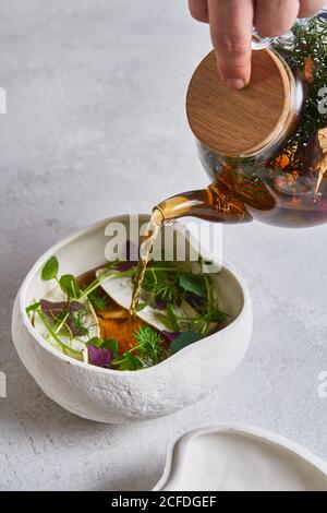 unrecognizable person pouring fresh warm tea from glass teapot into ceramic bowl with herbs Stock Photo