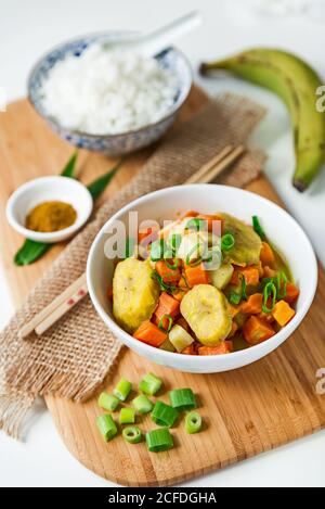 Asian curry with plantain and a bowl of white rice on wooden board with chopsticks and decoration Stock Photo