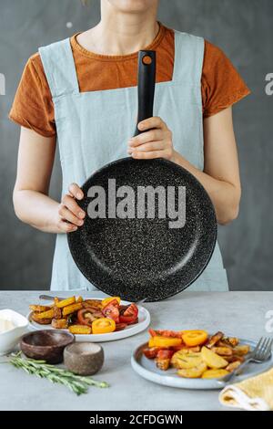 unrecognizable female with empty frying pan standing near table with delicious roasted vegetables in kitchen Stock Photo