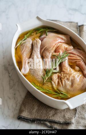 White ceramic baking dish with raw marinated chicken legs with sauce and fresh rosemary sprigs placed on cloth on marble table Stock Photo