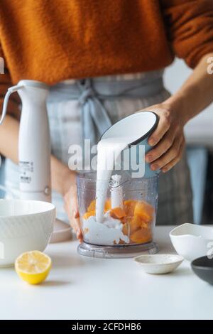 Crop cook carefully pouring milk to blender with pumpkin hand table with citrus in light kitchen Stock Photo