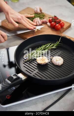 Grill pan with sprigs of fresh rosemary and cut garlic head placed on stove and crop hands of Woman crushing garlic on wooden cut board in background Stock Photo