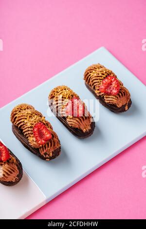 From above delectable chocolate eclairs with pieces of fresh raspberries and caramel cream placed in line on board on pink background Stock Photo