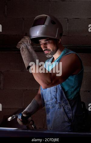 Professional man with welding mask on head wearing jeans overalls and protective gloves standing on workplace and preparing instrument for soldering while looking away