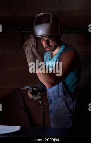 Professional man with welding mask on head wearing jeans overalls and protective gloves standing on workplace and preparing instrument for soldering while looking at camera Stock Photo
