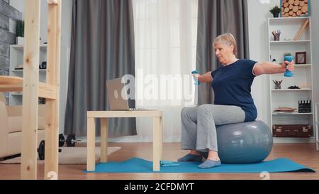 Healthy senior woman exercising with dumbbells in living room. Old person online internet training at home sport healthy lifestyle, elderly fitness exercise workout in apartment, activity and healthcare Stock Photo