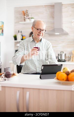 Elderly man surfing on internet using tablet computer during breakfast in kitchen. Senior person with tablet portable pad PC in retirement age using mobile apps, modern internet online information technology with touchscreen Stock Photo