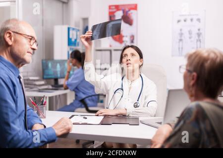 Female doctor looking at senior patient x-ray during consultation after surgery. Medic examining radiography in hospital office. Nurse working on computer. Stock Photo