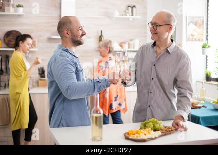 Happy son and father clinking wine glasses in kitchen during lunch with family. Stock Photo