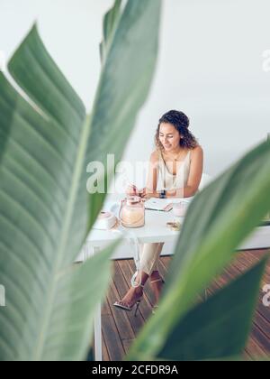 Cute young lady smiling and making notes in notepad while sitting at table in stylish office Stock Photo